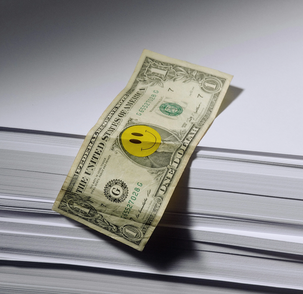 A dollar bill with a yellow smiley face sticker on it, placed on top of a large stack of white paper.